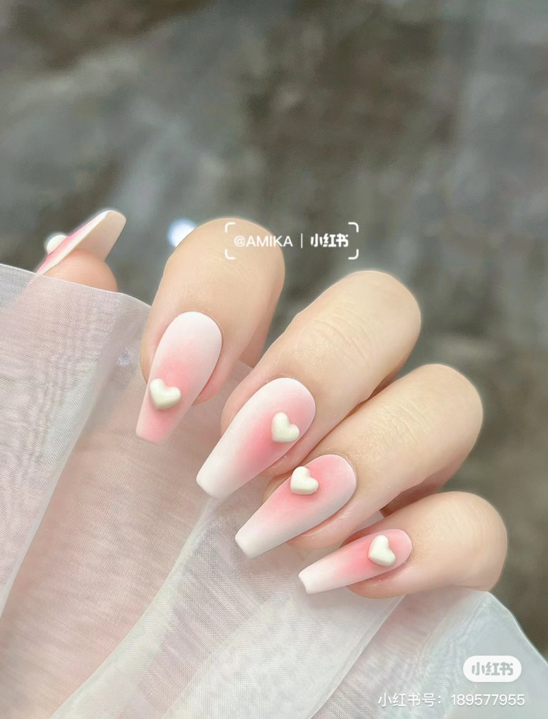Bàn tay với móng tay dài, sơn ombre hồng trắng, trang trí hình trái tim nhỏ trên nền vải mỏng.