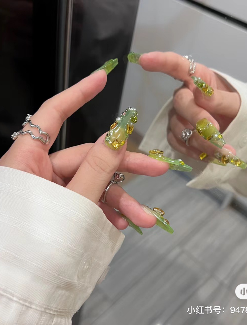 Bàn tay với móng tay dài màu xanh lá, trang trí đá và nhẫn bạc, chụp trước gương.