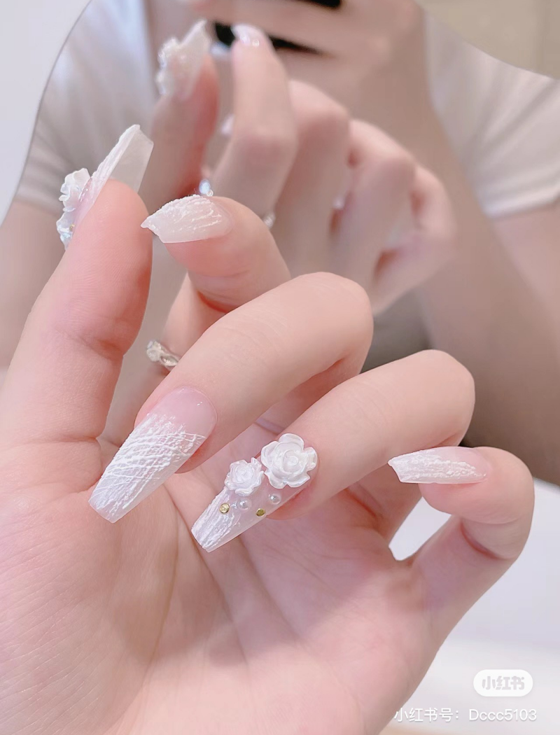 Bàn tay với móng tay trang trí hoa nổi và hạt ngọc, được chụp cận cảnh trước gương.