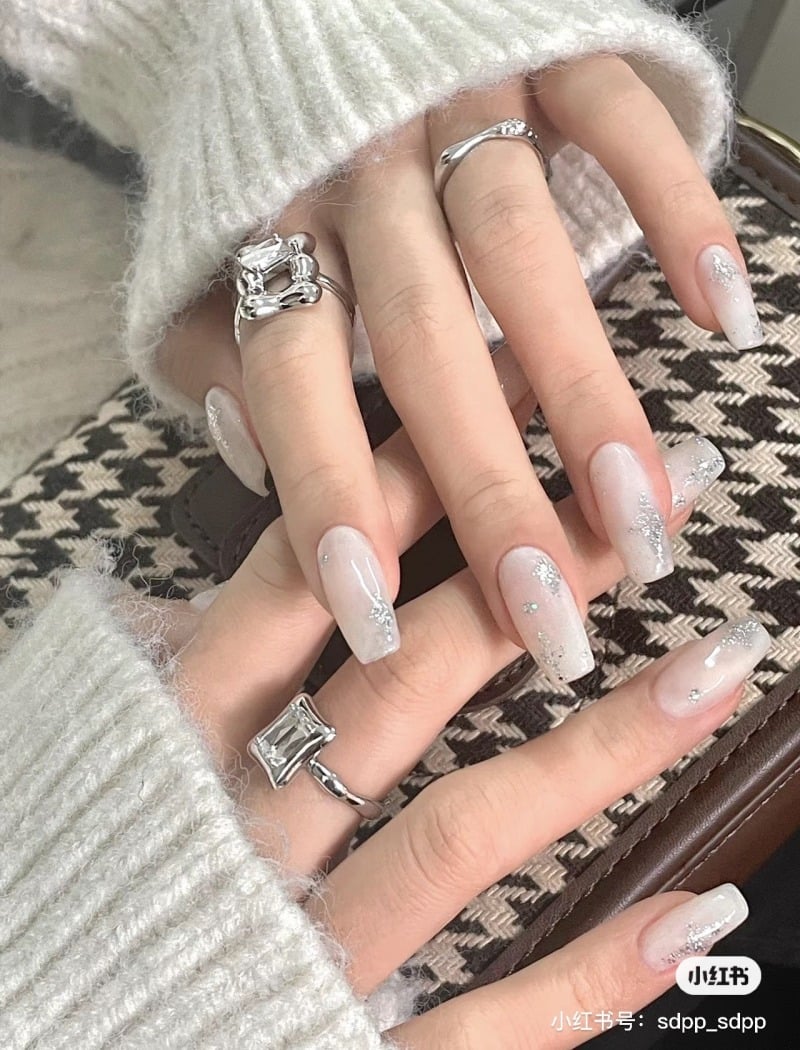 Bàn tay đeo nhẫn bạc, móng tay sơn màu nhạt, trang trí kim tuyến, áo len trắng, nền vải họa tiết.