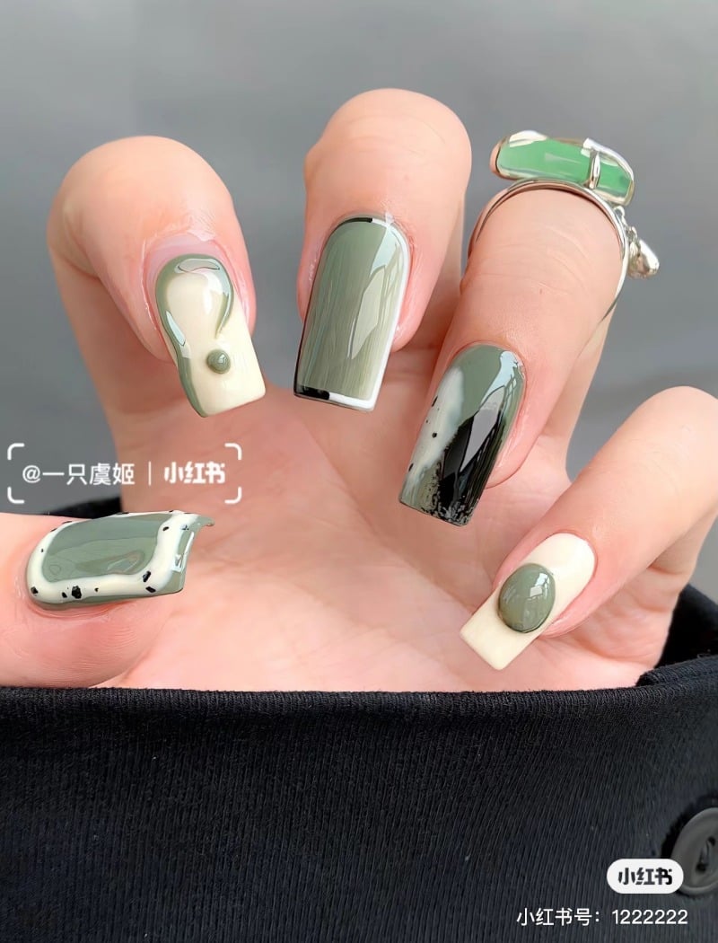 Bàn tay với móng tay có nghệ thuật sơn màu xanh lá và trắng, kèm nhẫn bạc trên ngón tay trỏ.