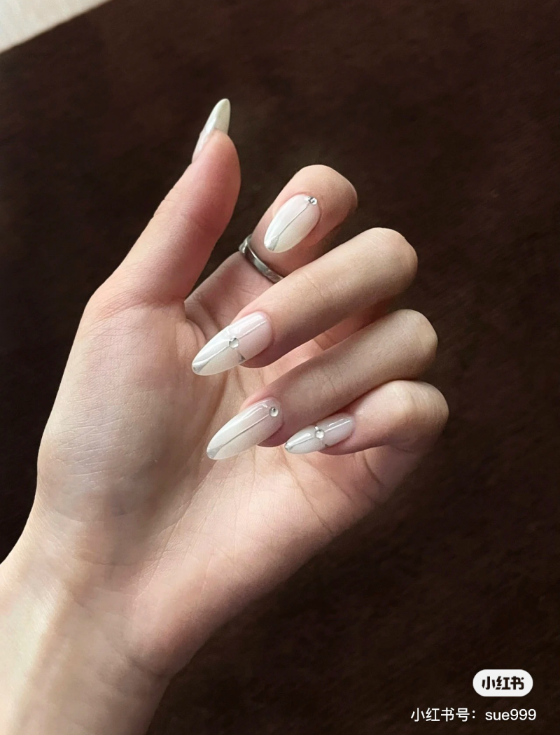 Bàn tay với móng tay sơn màu trắng, thiết kế đính đá nhỏ, trên nền màu nâu tối.