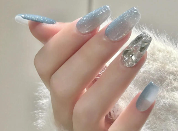 Tổng Hợp Mẫu Nail Màu Xanh Dương Nhạt Đẹp Nhẹ Nhàng Hot Nhất 2025 1 Bàn tay với móng tay dài được sơn màu xanh ánh kim loại, một móng tay đính đá lấp lánh, nền trắng mờ.