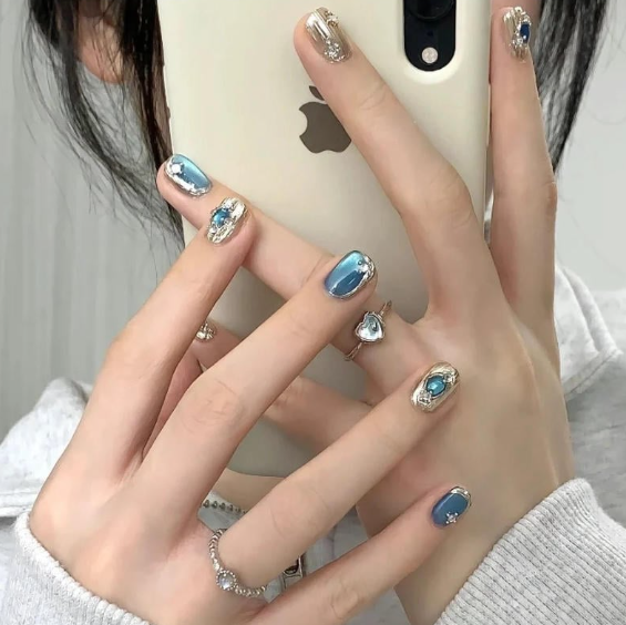 Tổng Hợp Mẫu Nail Màu Xanh Dương Nhạt Đẹp Nhẹ Nhàng Hot Nhất 2025 2 Tay cầm điện thoại, móng tay sơn màu xanh và đính đá, đeo nhiều nhẫn bạc, mặc áo xám nhạt.