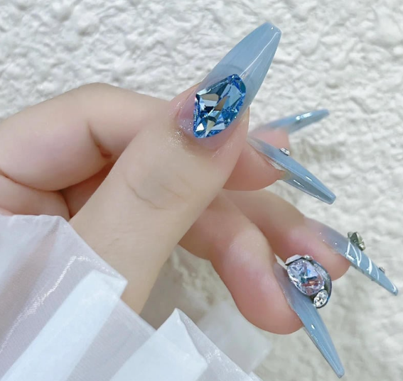 Tổng Hợp Mẫu Nail Màu Xanh Dương Nhạt Đẹp Nhẹ Nhàng Hot Nhất 2025 16 Bàn tay với móng tay dài màu xanh, trang trí đá quý màu xanh và ánh bạc, nền tường trắng.