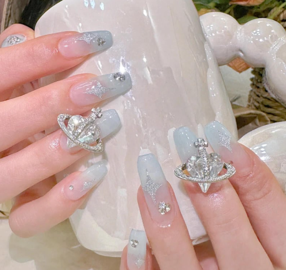 Tổng Hợp Mẫu Nail Màu Xanh Dương Nhạt Đẹp Nhẹ Nhàng Hot Nhất 2025 17 Bàn tay với móng tay dài, trang trí lấp lánh và phụ kiện hình hành tinh, cầm một chiếc cốc sứ trắng.