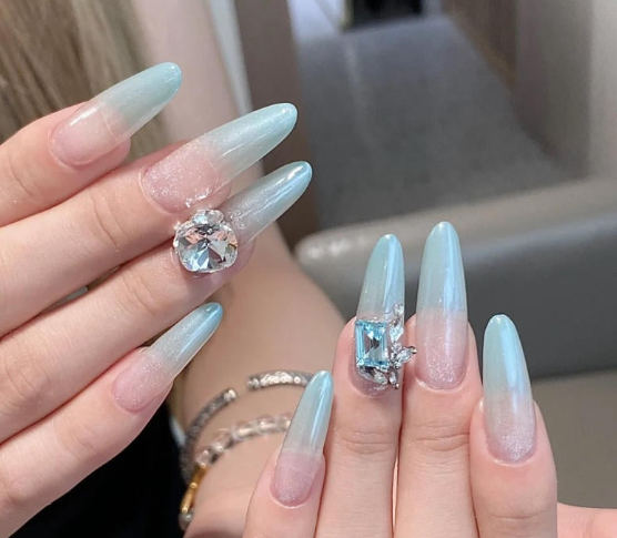 Tổng Hợp Mẫu Nail Màu Xanh Dương Nhạt Đẹp Nhẹ Nhàng Hot Nhất 2025 19 Bàn tay với móng tay dài, sơn màu xanh nhạt, đính đá lớn lấp lánh, nền nhạt phía sau.