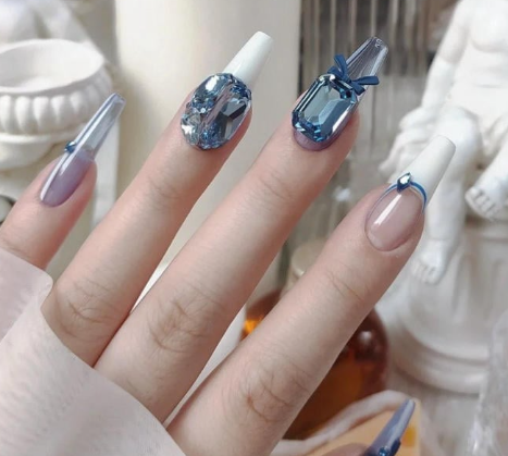 Tổng Hợp Mẫu Nail Màu Xanh Dương Nhạt Đẹp Nhẹ Nhàng Hot Nhất 2025 20 Bàn tay với móng tay dài, trang trí đá màu xanh và trắng, nền là các vật dụng trang trí màu trắng.