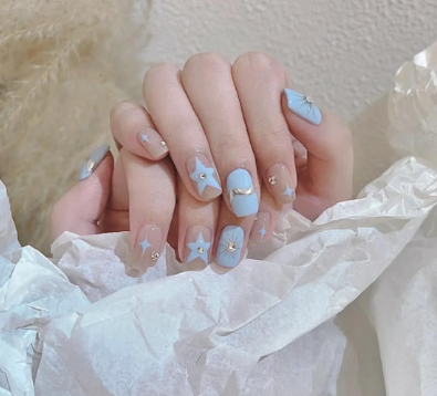 Tổng Hợp Mẫu Nail Màu Xanh Dương Nhạt Đẹp Nhẹ Nhàng Hot Nhất 2025 28 Bàn tay với móng tay được sơn màu xanh nhạt, trang trí hình ngôi sao và mặt trăng, trên nền giấy trắng.