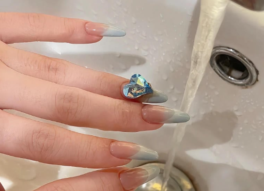 Tổng Hợp Mẫu Nail Màu Xanh Dương Nhạt Đẹp Nhẹ Nhàng Hot Nhất 2025 33 Ngón tay với móng nghệ thuật dài, đính đá hình trái tim, dưới vòi nước đang chảy trong bồn rửa.