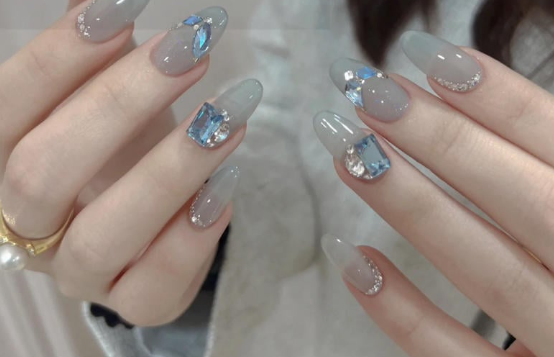 Tổng Hợp Mẫu Nail Màu Xanh Dương Nhạt Đẹp Nhẹ Nhàng Hot Nhất 2025 39 Bàn tay với móng tay dài được sơn màu xám bóng, trang trí đá quý lấp lánh và nhẫn vàng trên ngón tay.