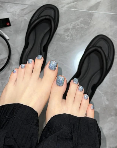 Tổng Hợp Mẫu Nail Màu Xanh Dương Nhạt Đẹp Nhẹ Nhàng Hot Nhất 2025 67 Bàn chân với móng chân sơn màu lấp lánh, đặt trên sàn đá, cạnh đôi dép màu đen.