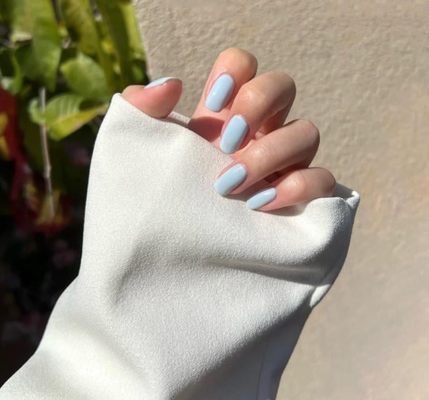 Tổng Hợp Mẫu Nail Màu Xanh Dương Nhạt Đẹp Nhẹ Nhàng Hot Nhất 2025 77 Tay với móng tay sơn màu xanh nhạt, áo tay trắng, nền tường sáng và cây xanh phía xa.