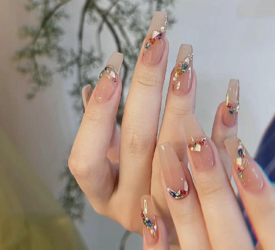 99+ mẫu nail đính đá vừa XINH vừa SANG cho nàng tự tin ra ngoài 14 Tay với móng tay dài, sơn màu nude và đính đá màu sắc, nền có cây trang trí.
