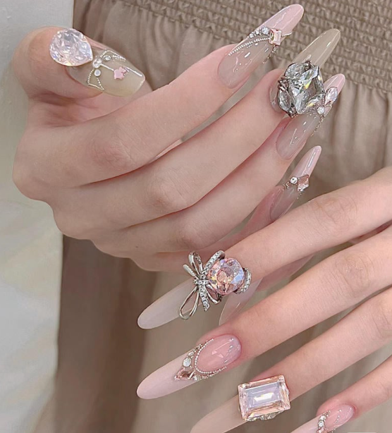 99+ mẫu nail đính đá vừa XINH vừa SANG cho nàng tự tin ra ngoài 21 Bàn tay với móng tay dài, trang trí đá quý và phụ kiện lấp lánh, trên nền váy màu nâu nhạt.