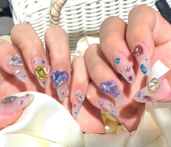 99+ mẫu nail đính đá vừa XINH vừa SANG cho nàng tự tin ra ngoài 22 Bàn tay với móng tay dài đính các viên đá trang trí màu sắc khác nhau, đặt trên nền vải sáng màu.