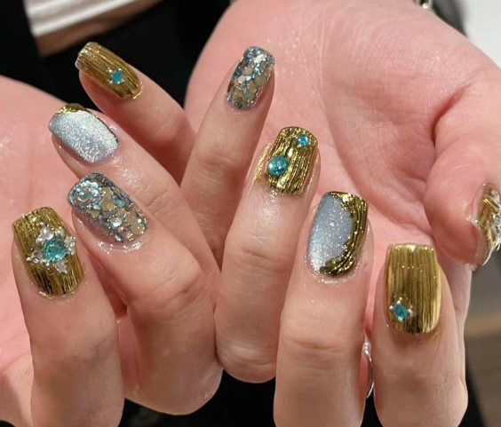 99+ mẫu nail đính đá vừa XINH vừa SANG cho nàng tự tin ra ngoài 23 Bàn tay với móng tay được trang trí bằng sơn màu vàng và bạc kèm họa tiết đính đá xanh.