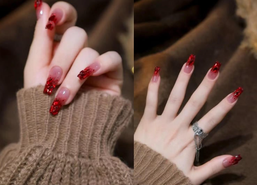 99+ mẫu nail đính đá vừa XINH vừa SANG cho nàng tự tin ra ngoài 32 Bàn tay với móng tay dài sơn đỏ lấp lánh, một tay đeo nhẫn, áo len màu nâu.