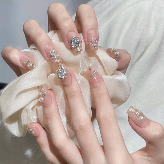 99+ mẫu nail đính đá vừa XINH vừa SANG cho nàng tự tin ra ngoài 48 Bàn tay với móng tay đính đá và ngọc trai, cầm một chiếc dây buộc tóc vải satin màu trắng.