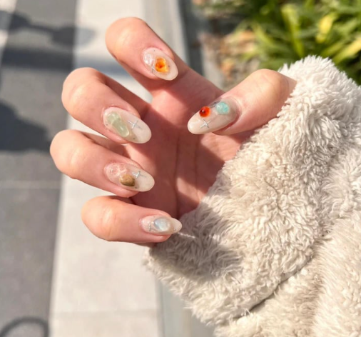 99+ mẫu nail đính đá vừa XINH vừa SANG cho nàng tự tin ra ngoài 53 Bàn tay với móng tay được trang trí bằng đá màu nổi bật, áo lông xám, nền là đường phố và cây xanh.