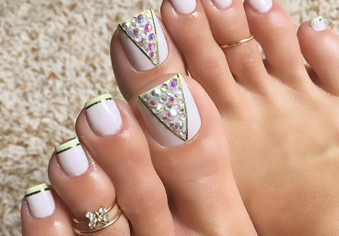 99+ mẫu nail đính đá vừa XINH vừa SANG cho nàng tự tin ra ngoài 64 Bàn chân với móng được sơn màu trắng, trang trí kim tuyến và nhẫn vàng, trên nền thảm màu sáng.