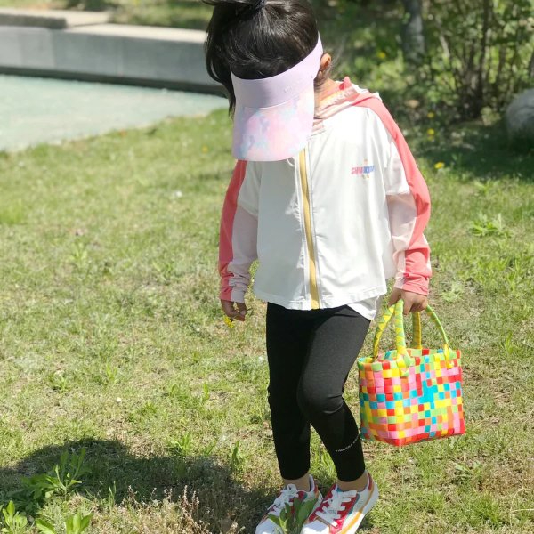 Bé gái đội mũ lưỡi trai màu hồng, mặc áo khoác trắng, cầm giỏ nhiều màu đứng trên bãi cỏ xanh.