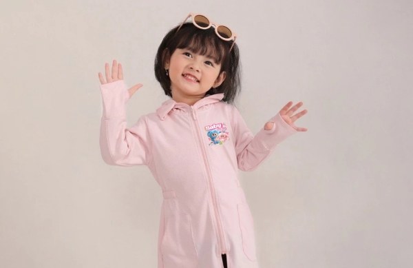 Bé gái mặc áo khoác hồng, đeo kính râm trên đầu, giơ tay tạo dáng vui vẻ trước nền màu trơn.