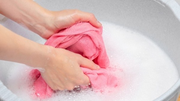 Đôi tay giặt áo len màu hồng trong chậu nước xà phòng trắng, tạo nhiều bọt.