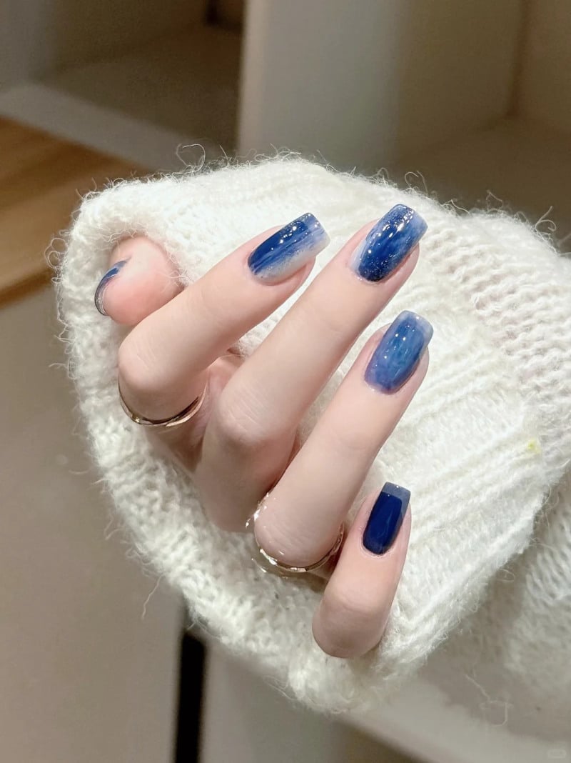 Bàn tay đeo nhẫn, móng tay sơn màu xanh lam ombre lấp lánh, áo len trắng.