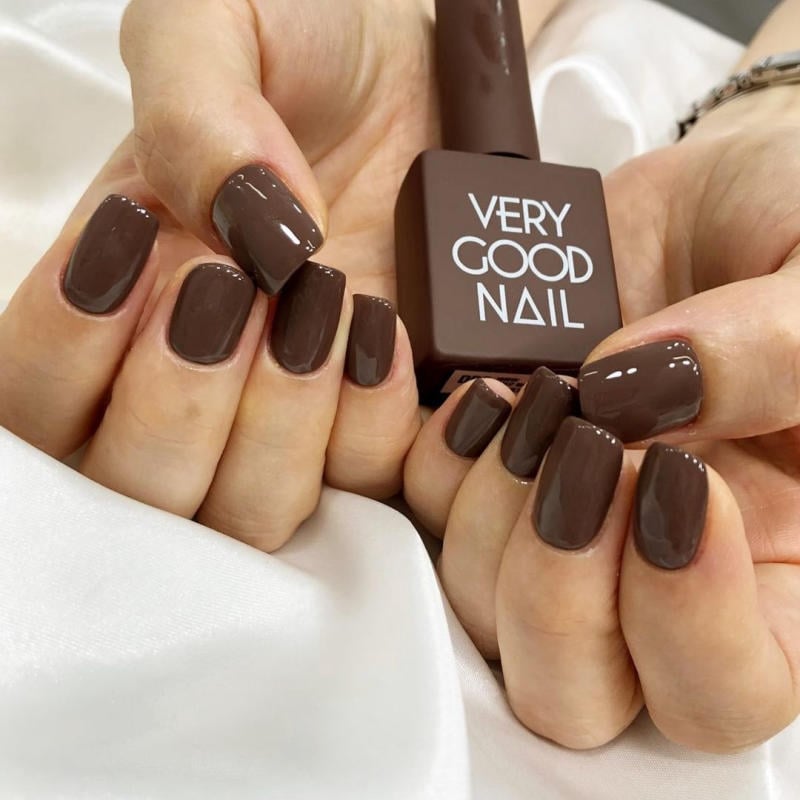 bang-mau-very-good-nail__1__1a10400ca2ff4e9a8d7de5e412b0432e Bàn tay với móng tay sơn màu nâu bóng, cầm lọ sơn có nhãn "Very Good Nail", nền vải trắng.