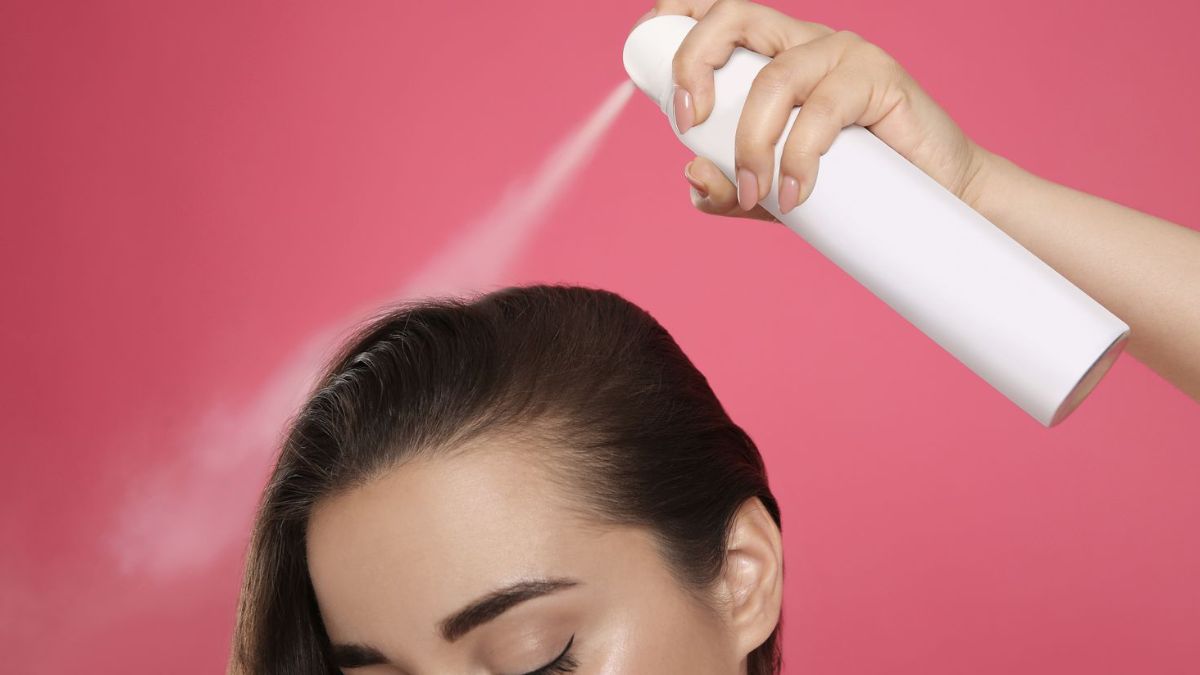 Người đang xịt keo lên tóc bằng chai xịt màu trắng, nền hồng.