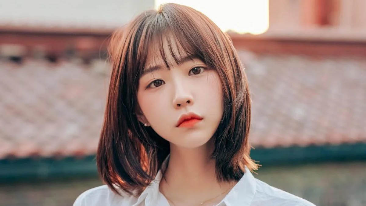 Người có mái tóc ngắn, mặc áo trắng, đứng trước nền trời sáng.
