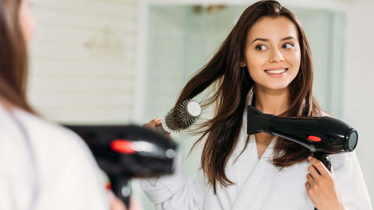 Người phụ nữ trong áo choàng tắm đang sấy tóc và chải tóc trước gương trong một phòng tắm sáng sủa.