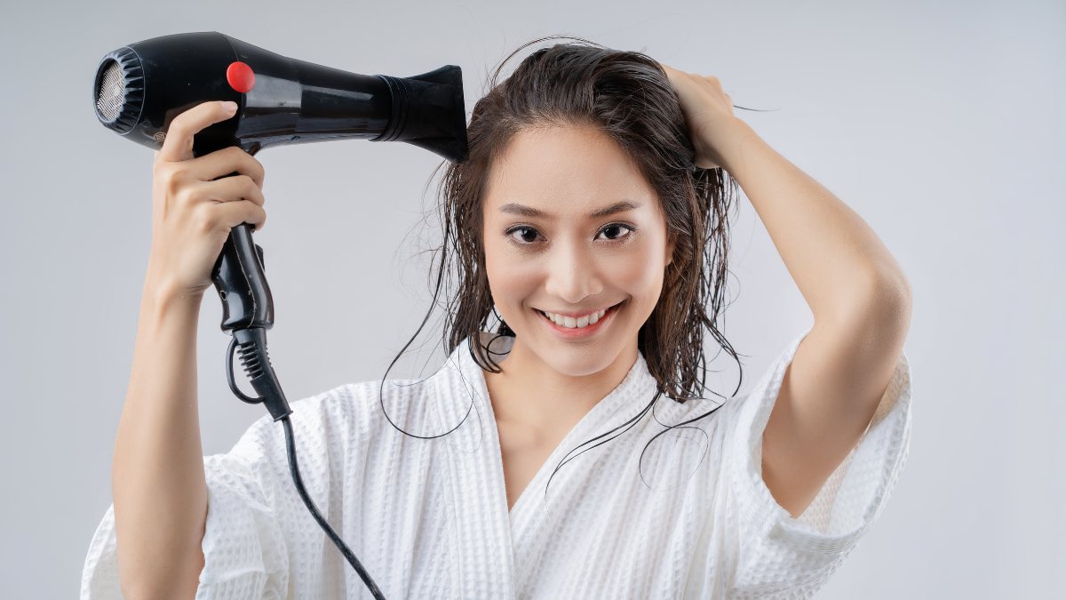 Người mặc áo choàng trắng đang sấy tóc bằng máy sấy cầm tay, tóc còn ướt và gương mặt tươi cười.