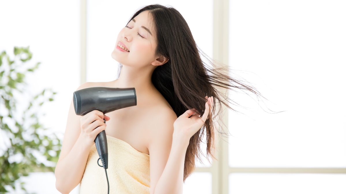 Người phụ nữ quấn khăn tắm, dùng máy sấy tóc, tóc bay nhẹ, phông nền cửa sổ sáng.