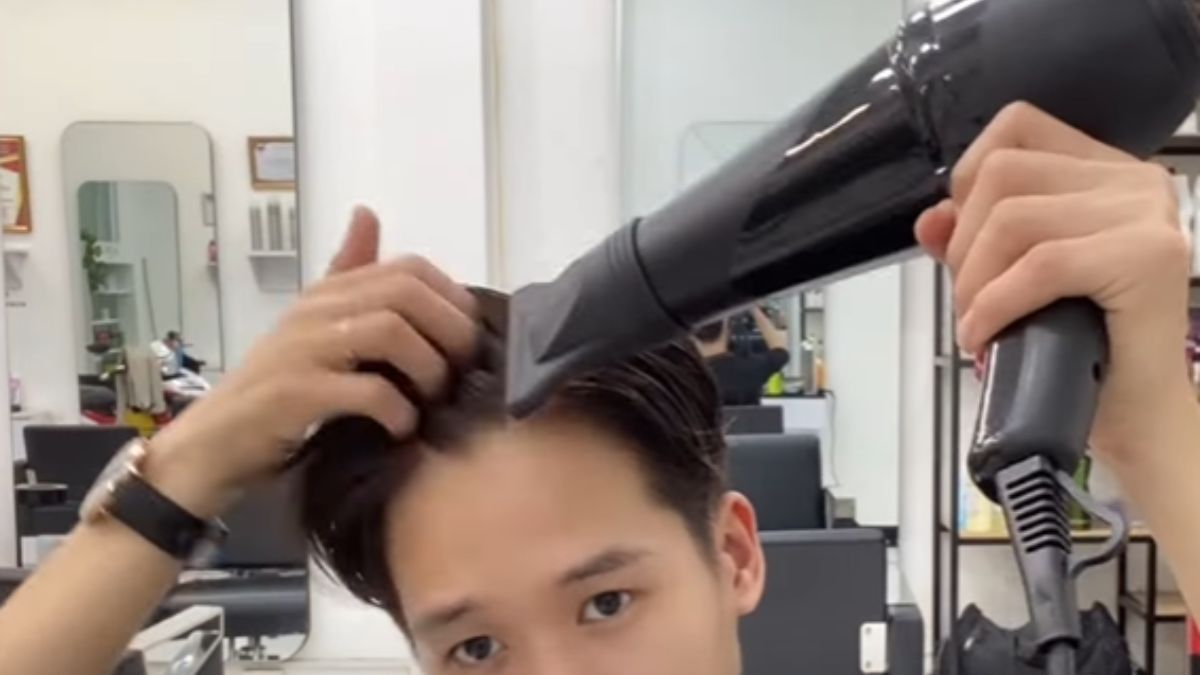 Người đang sấy tóc bằng máy sấy trong tiệm cắt tóc, xung quanh có gương và ghế cắt tóc.