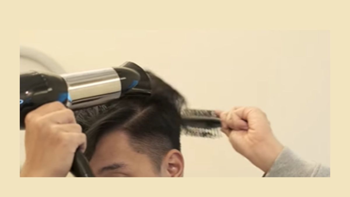 Người đang sấy tóc bằng máy sấy và lược tròn, tạo kiểu tóc trên nền tường màu nhạt.