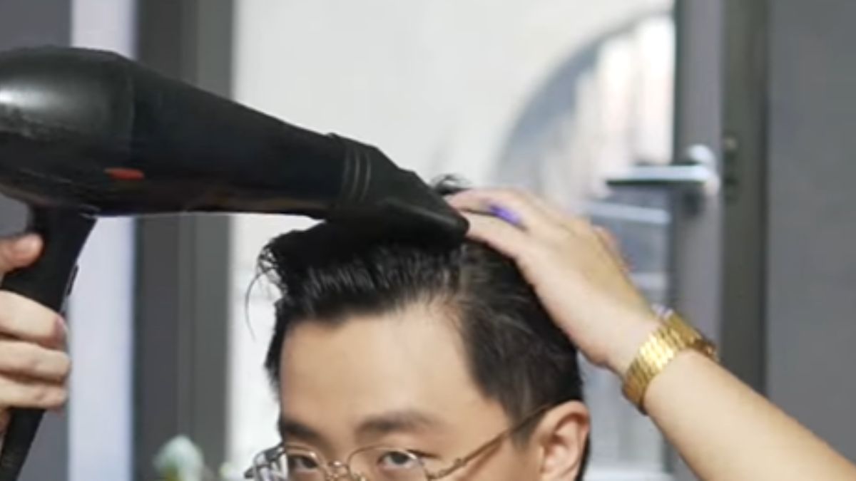 Người đang dùng máy sấy tóc để tạo kiểu tóc, tay còn lại vuốt nhẹ trên đầu.