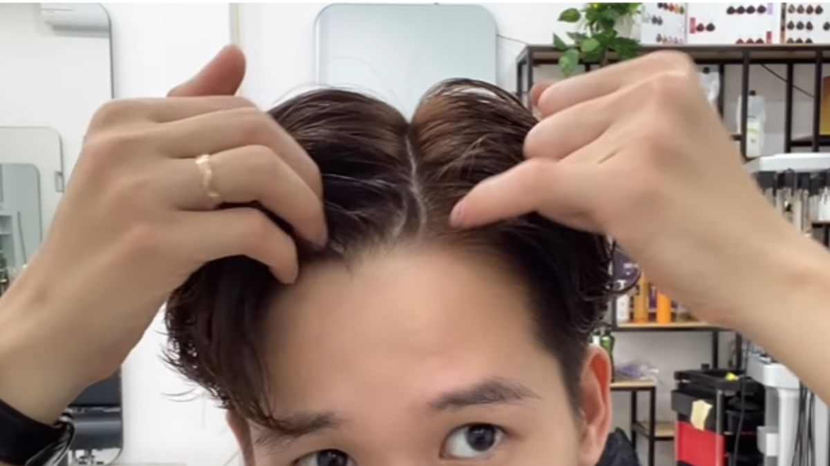 Người đang tự chia tóc giữa, tay chạm vào tóc, trong một tiệm làm tóc với kệ và gương.