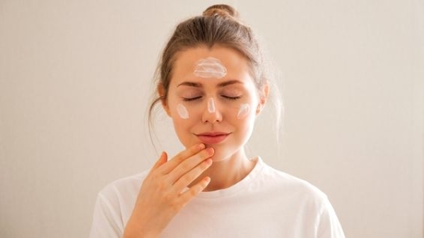 Người mặc áo trắng thoa kem dưỡng da lên mặt, mắt nhắm, tay chạm cằm, đứng trước nền trắng.
