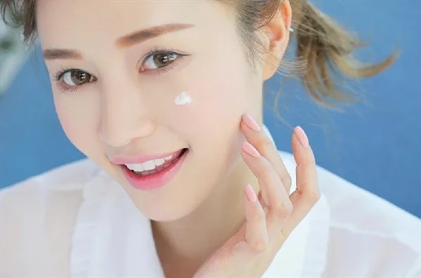 image20-16 Người phụ nữ thoa kem dưỡng da lên má, mỉm cười, mặc áo trắng, nền xanh.