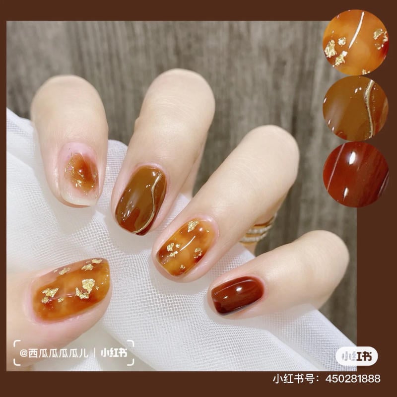 Bàn tay với móng tay được sơn màu nâu và cam nhạt, trang trí với hoạ tiết ánh kim, nền vải trắng.