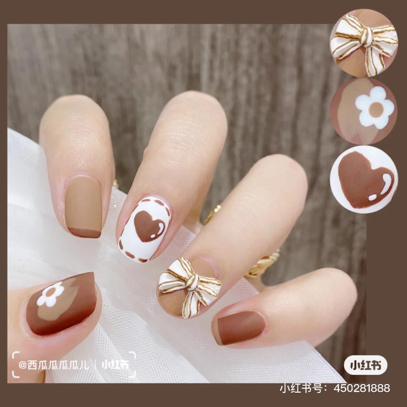 Bàn tay với móng tay được trang trí họa tiết nơ, hoa và trái tim trên nền màu nâu và trắng.