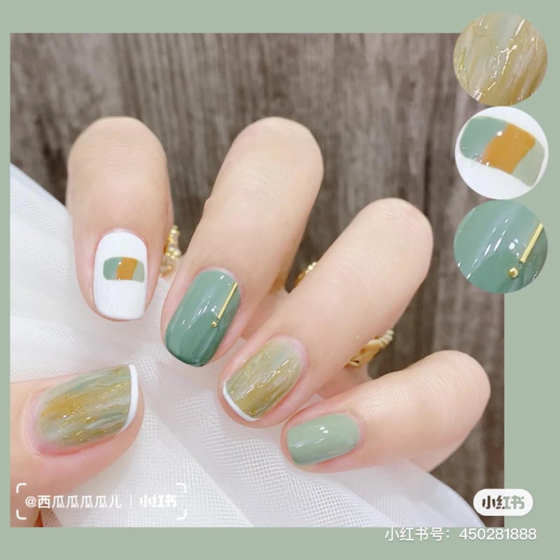 Bàn tay với móng tay được sơn nghệ thuật, kết hợp màu xanh lá, trắng và vàng, trên nền vải trắng.