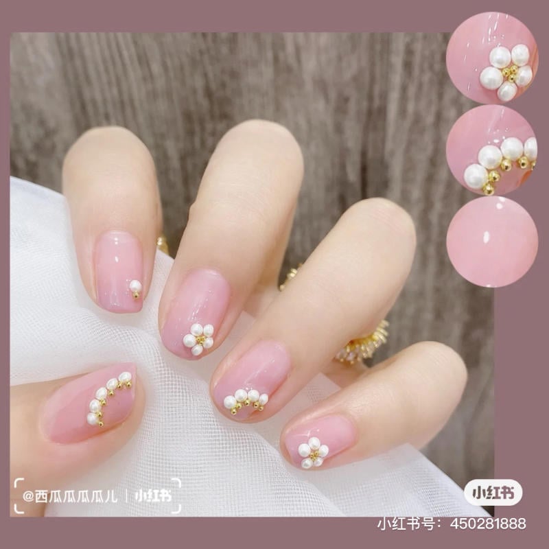 Bàn tay với móng tay màu hồng nhạt, trang trí hoa và hạt cườm trắng vàng, trên nền vải màu trắng.