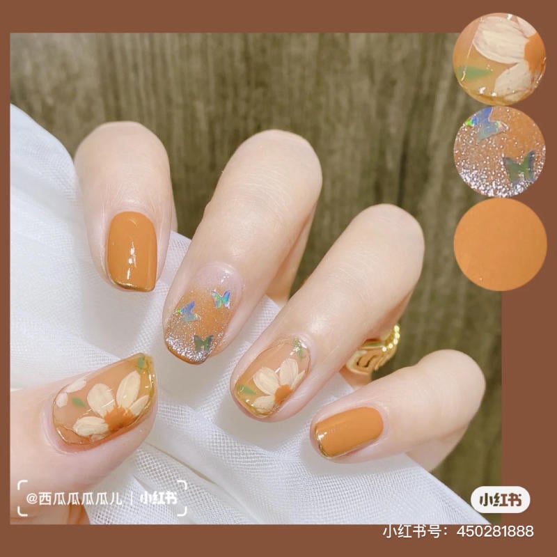 Bàn tay với móng tay sơn màu cam, trang trí họa tiết hoa và cánh bướm ánh kim.