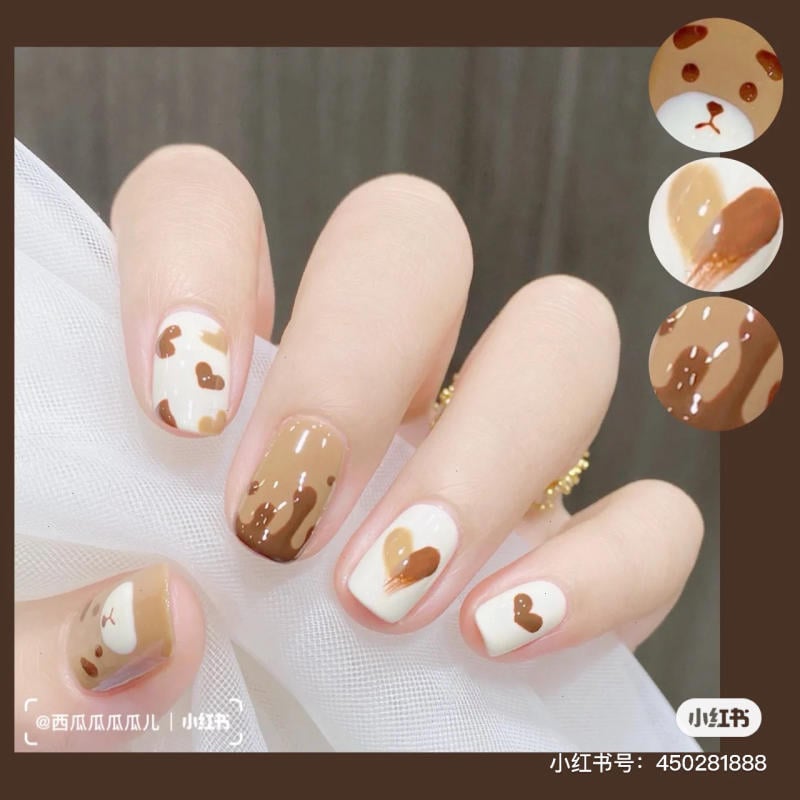 Bàn tay với móng tay được sơn nghệ thuật họa tiết gấu và trái tim màu nâu trên nền trắng và nâu.