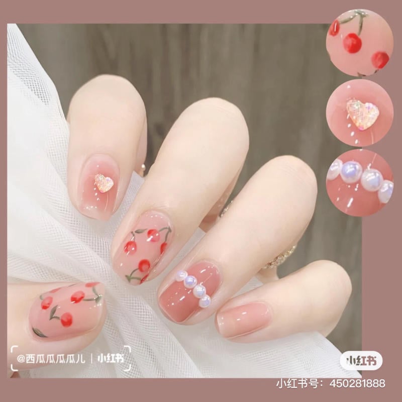 Bàn tay với móng tay được trang trí họa tiết trái cây và ngọc trai, tông màu hồng nhạt và đỏ nổi bật.