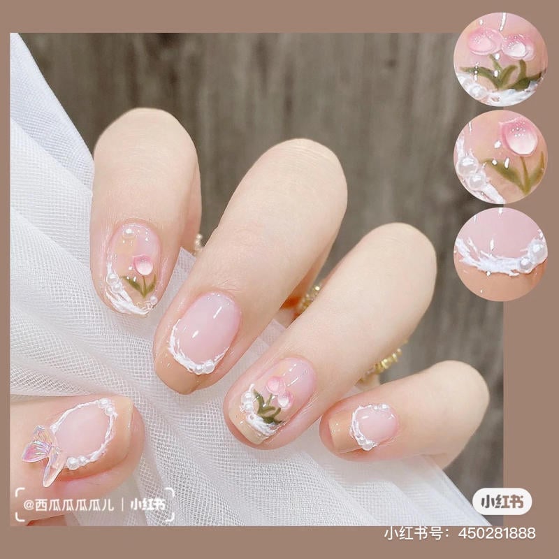 Bàn tay với móng đính hoa hồng và phụ kiện trang trí tinh tế trên nền màu hồng nhạt.