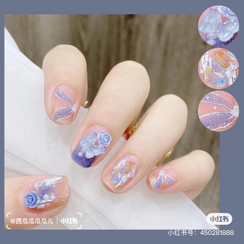 Bàn tay với móng tay được trang trí họa tiết xanh lam, hoa và ánh kim loại lấp lánh trên nền màu tự nhiên.